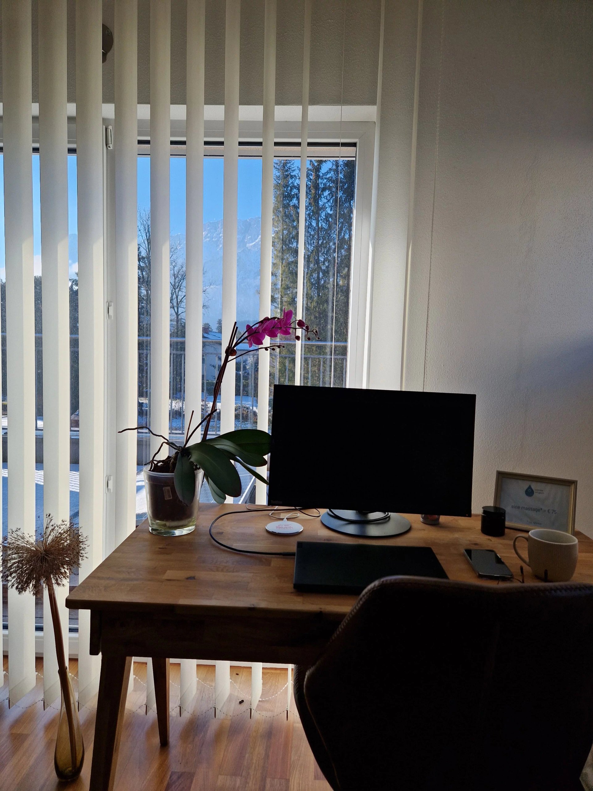 Ein Schreibtisch mit Laptop, Tasse, Handy und einer Orchidee steht vor einem Fenster mit Lamellen und Blick auf Berge, das Bild repräsentiert die Arbeit in der IT.